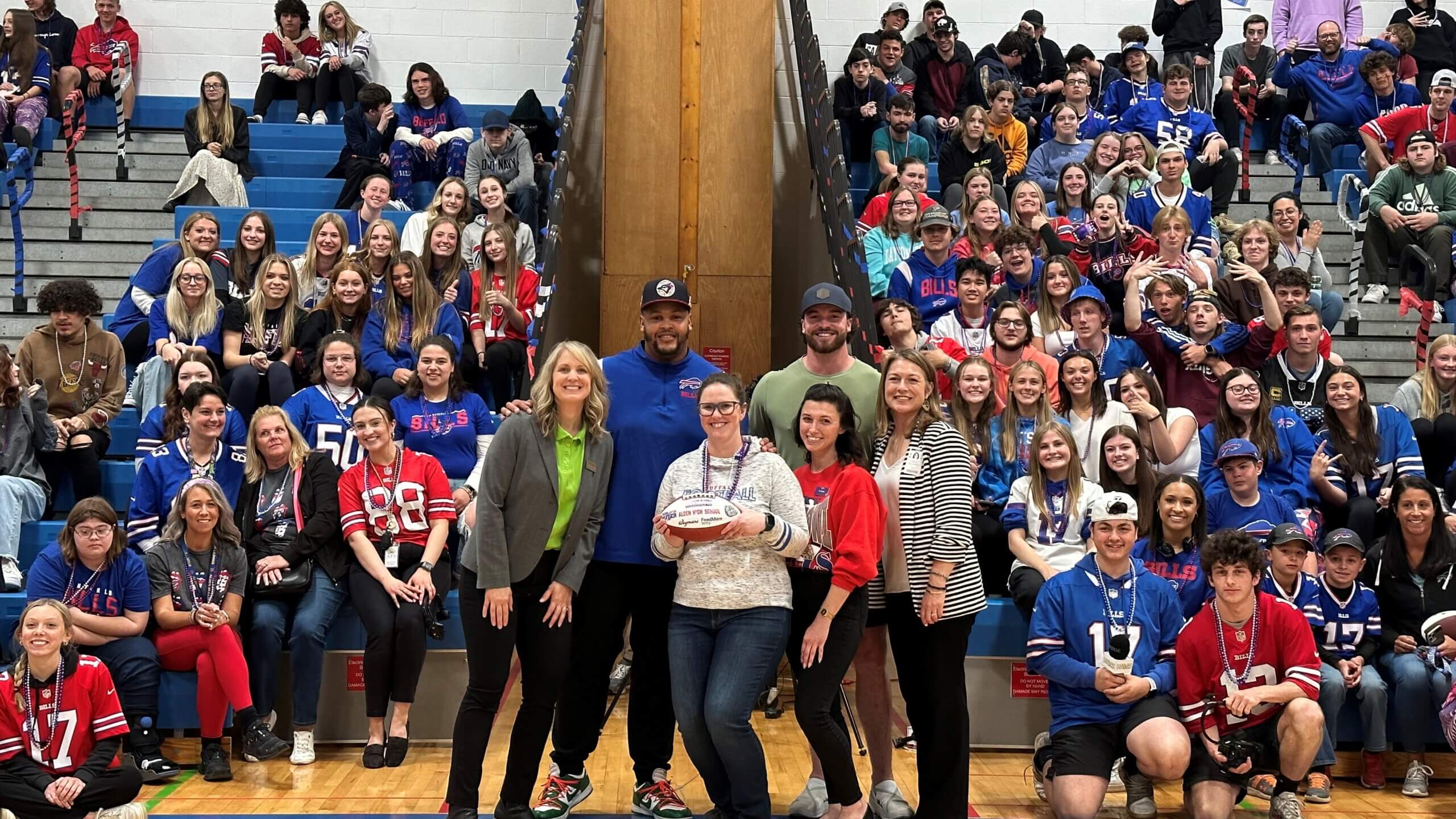 Buffalo Bills players Dion Dawkins and Dawson Knox, representatives from Wegmans and the Bills Foundation, and high school staff members hold football in front of gymnasium stands filled with high school students.